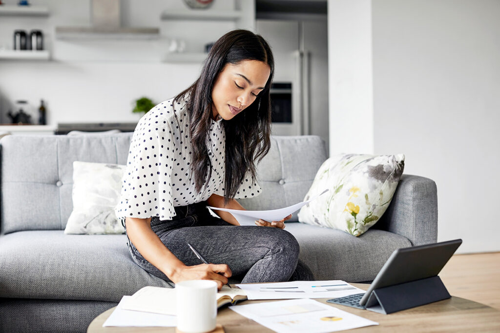 Woman in polka dot white shirt sitting on a couch holding papers and looking at a laptop computer.