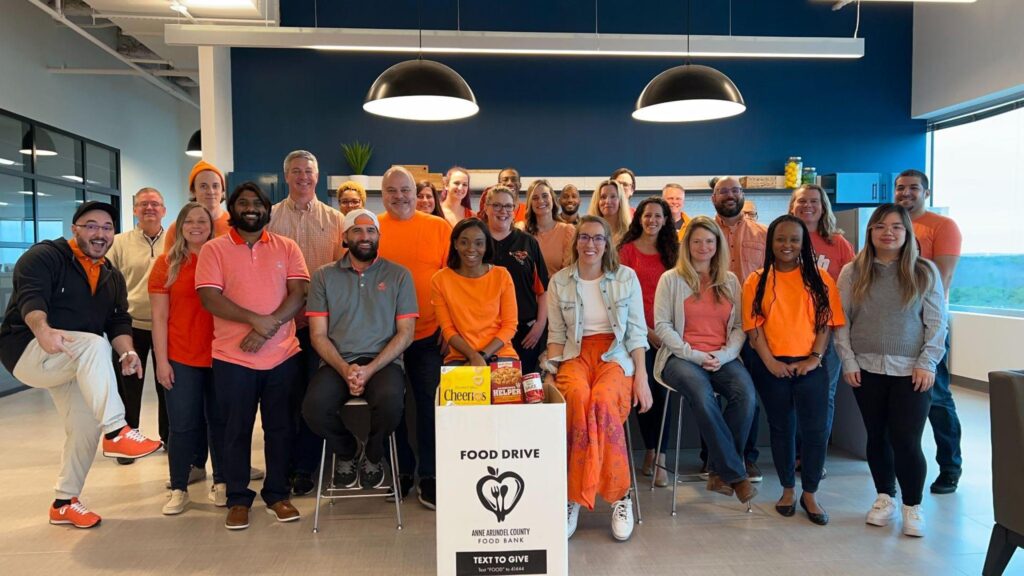 CallTrackingMetrics' employees pose in orange-colored attire with a large box of food donations they collected for Anne Arundel County Food Bank. 